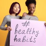 Two women holding a sign with '#healthy habits' on a vibrant yellow background.