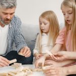 Four family members of different ages engage with toy blocks on a table indoors.